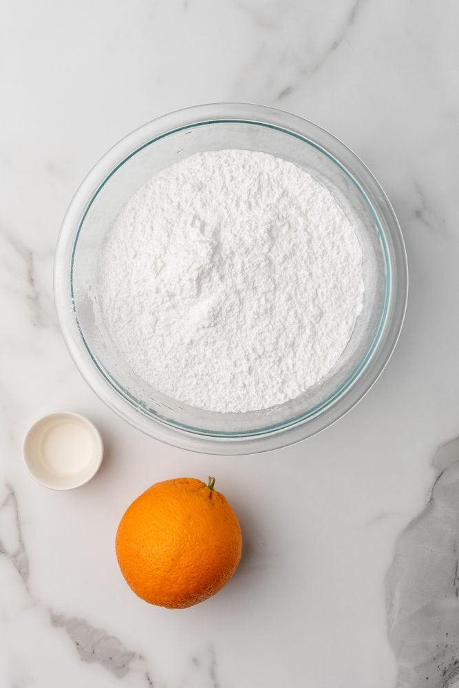 Three ingredients to make orange glaze cake icing: a large bowl of powdered sugar, a small bowl of orange extract, and one whole orange.