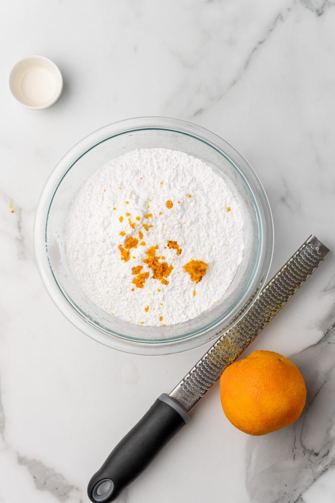 Large glass bowl of powdered sugar with fresh orange zest sprinkled in the middle, microplane grater and whole orange with peel zested off to the side.