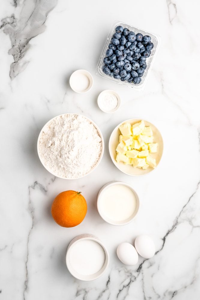 Overhead photo of ingredients for blueberry scone recipe including pint of blueberries, sugar, flour, salt, butter, cream, eggs, and whole orange.