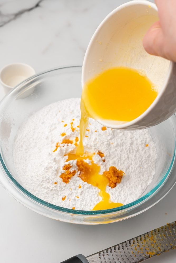 Pouring orange juice into glass bowl with powered sugar and orange zest.