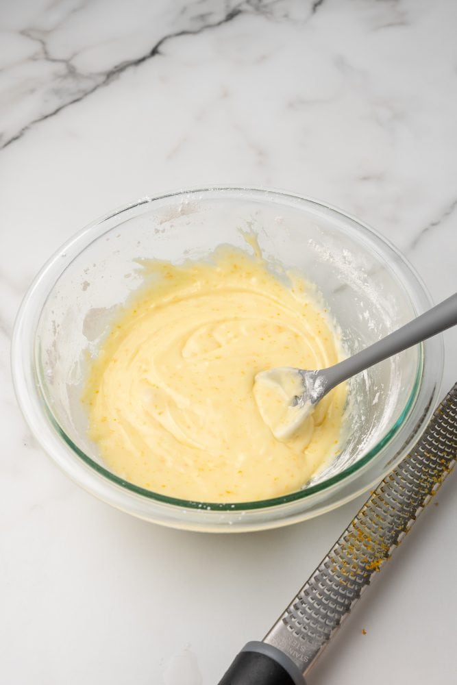 Mixing together powedered sugar, orange zest, orange juice and orange extract in a clear glass bowl. The consistency is of a light frosting and the color is light orange yellow with darker orange specks.