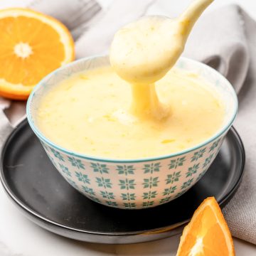 Orange glaze cake icing in a small serving bowl with a spoon taking some glaze off the top. There's a sliced orange in the background and an orange wedge in the foreground.