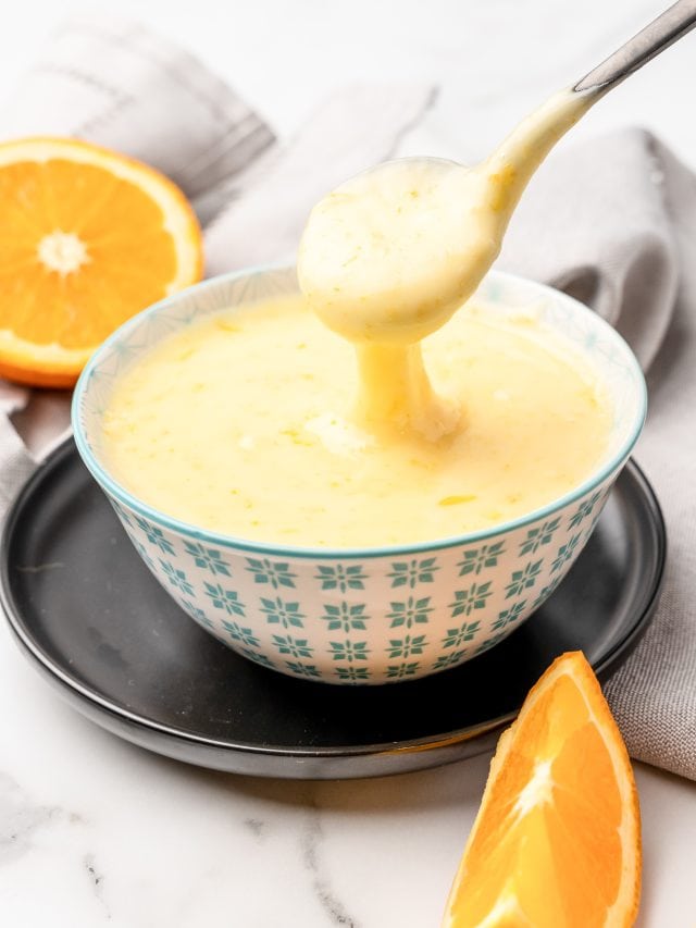 Orange glaze cake icing in a small serving bowl with a spoon taking some glaze off the top. There's a sliced orange in the background and an orange wedge in the foreground.