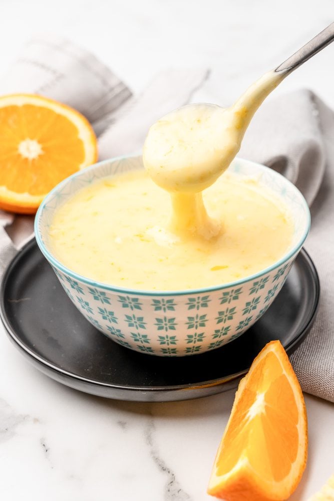 Orange glaze cake icing in a small serving bowl with a spoon taking some glaze off the top. There's a sliced orange in the background and an orange wedge in the foreground.