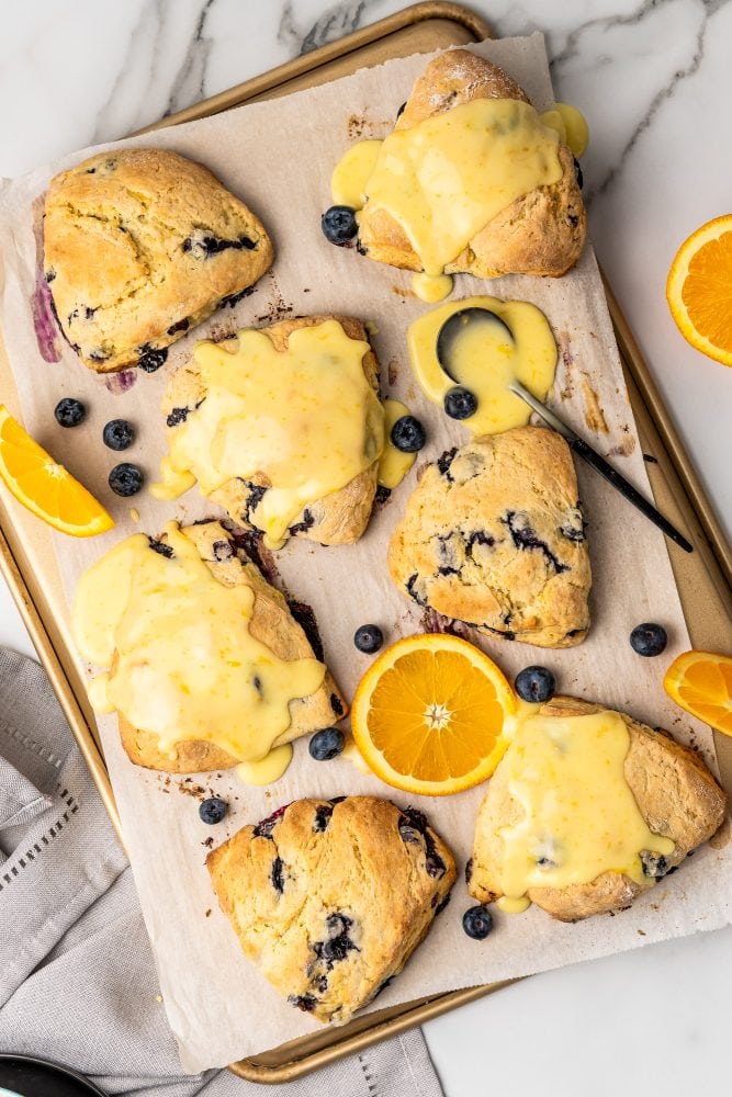 Overhead photo of a baking pan lined with parchment paper covered in blueberry scones with slices of orange and blueberries for garnish.
