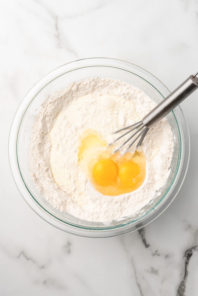 Clear mixing bowl with flour, heavy cream, and two eggs and a whisk.