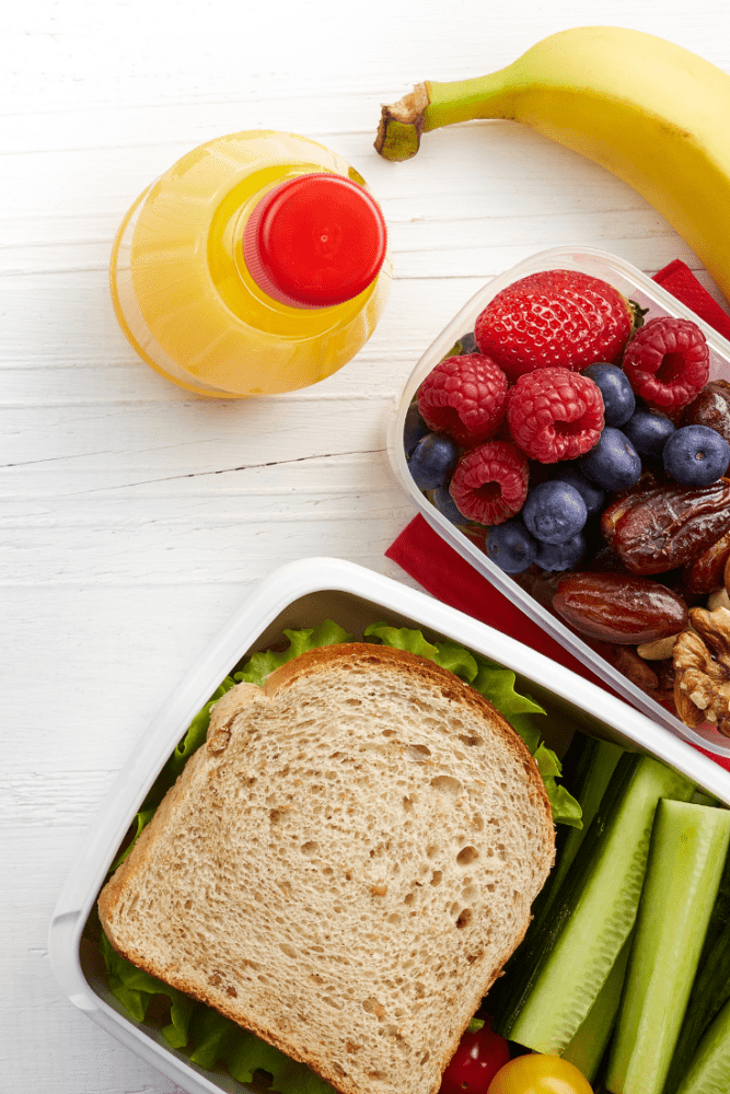 Photo of lunch box with sandwich, orange juice, mixed berries, dates, banana, and cucumber.