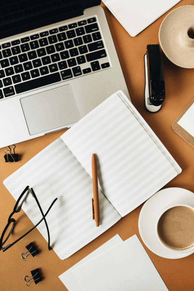 Overhead flatlay of desk with macbook, notebook opened with blank pages, pen, cup of coffee, stapler, glasses, and binder clips.