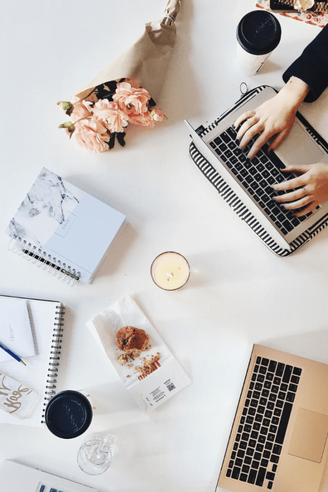 Overhead flatlay shot of at-home desk including two macbooks (a woman's hands typing on one), fresh flowers, planners, coffee, and a candle.