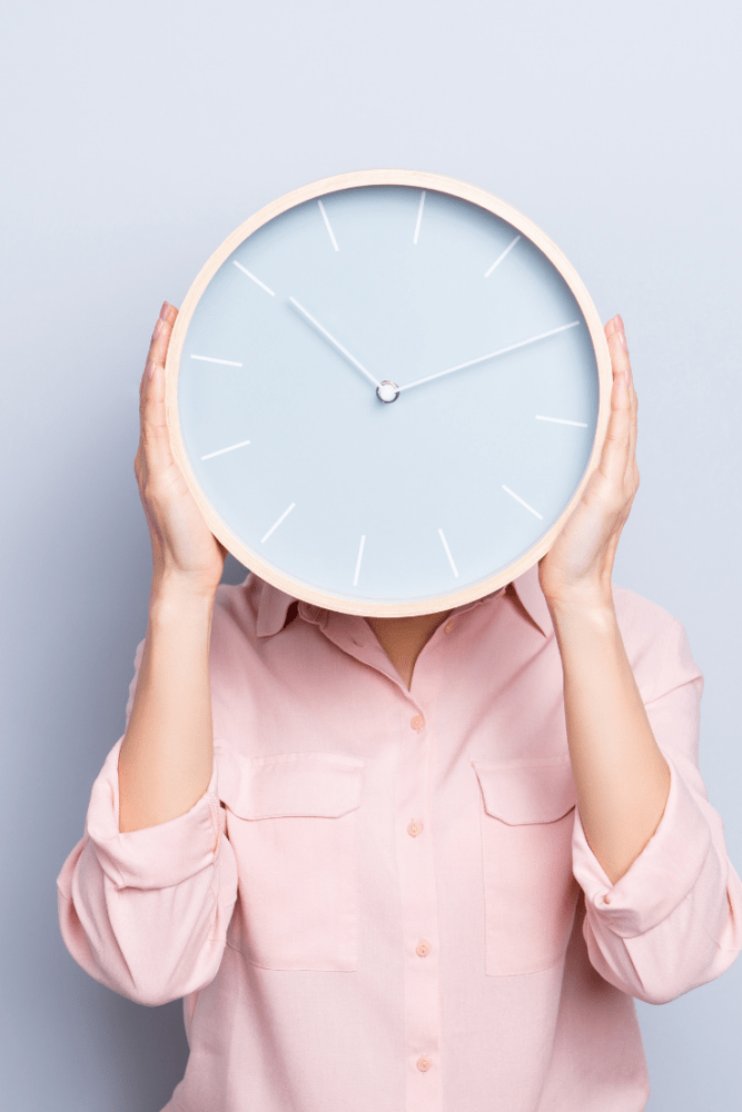 Woman wearing a pink button down shirt holding a big clock in front of her face.