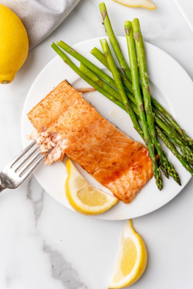 Serving of maple glazed sockeye salmon on a white plate with lemon slices and asparagus. A fork is taking a piece showing that the salmon flakes and is tender.