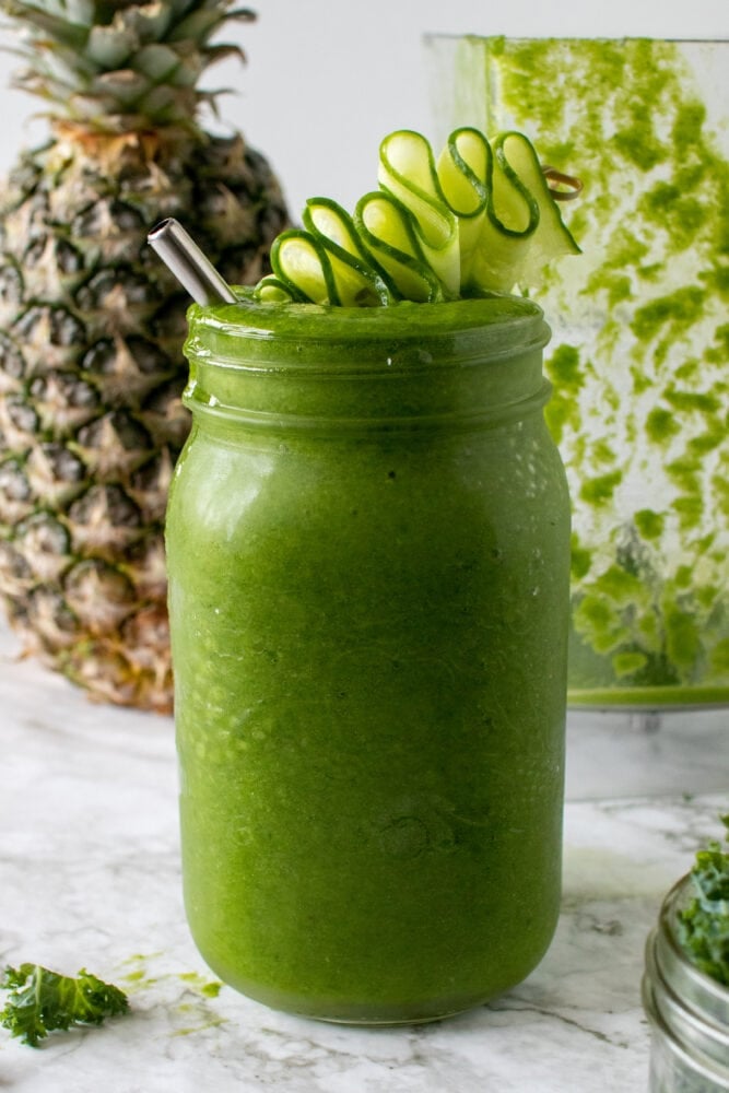 Large mason jar with pineapple kale green smoothie an a cucumber garnish on top.