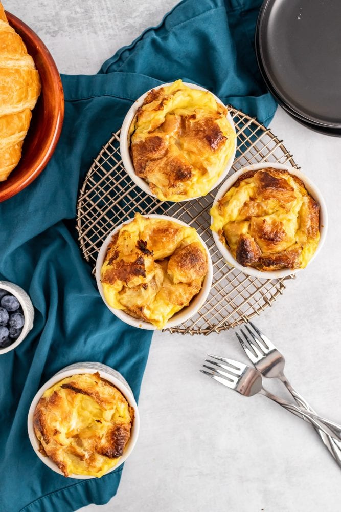 Three individual ramekins with baked croissant bread pudding cooling on a wire rack, with an additional bread pudding off to the side with forks ready to serve.