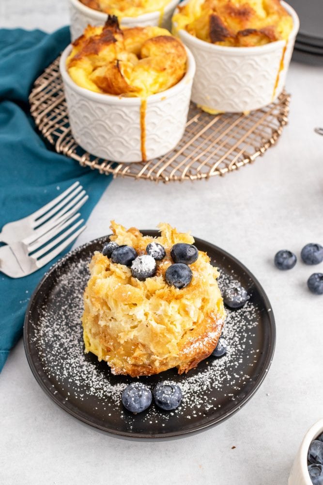 One croissant bread pudding served on a plate, topped with blueberries and powdered sugar.