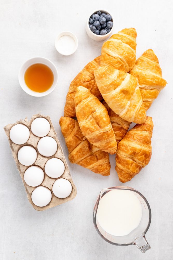 Overhead view of all the ingredients needed for croissant bread pudding including eggs, Grand Marnier, sugar, milk, croissants, and blueberries for garnish.