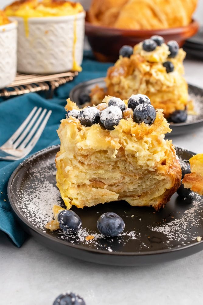 Side view of individual serving of croissant bread pudding showing the flaky layers of croissant, topped with blueberries and powdered sugar.