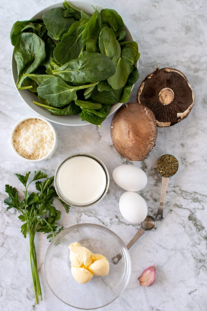 Overhead shot of ingredients to make baked portobello mushrooms: spinach, parmesan cheese, cream, garlic, parsley, eggs, mushrooms, and Italian seasoning.