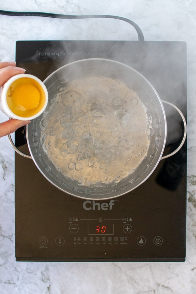 Overhead shot of sliding egg from a small bowl into a pot of boiling water.