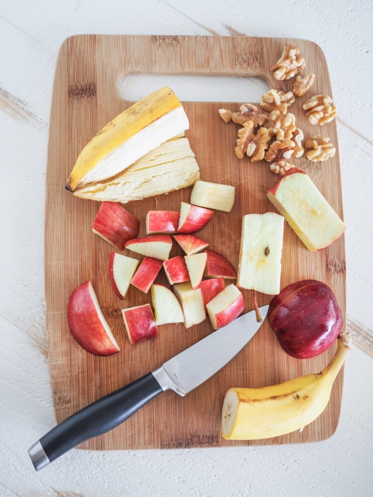 Bamboo cutting board showing some of the apple smoothie ingredients including half a banana, chopped apple, and walnuts.