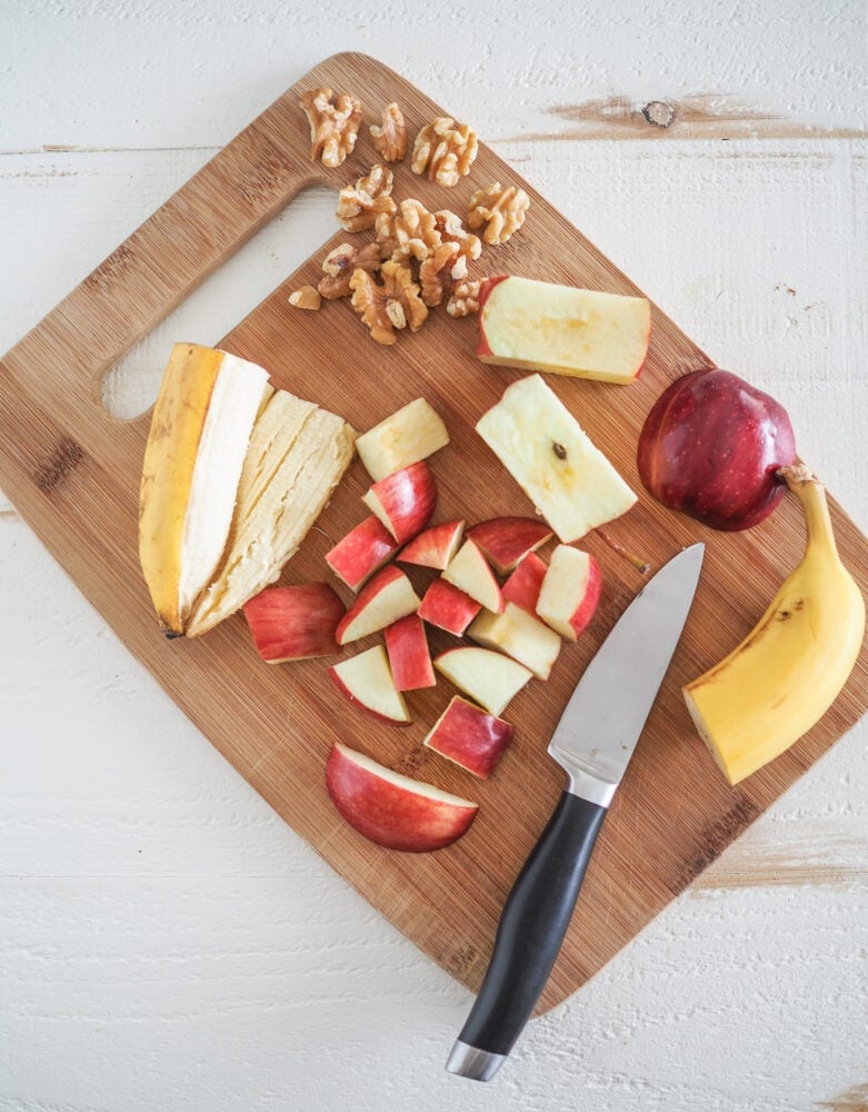 Bamboo cutting board showing some of the apple smoothie ingredients including half a banana, chopped apple, and walnuts.