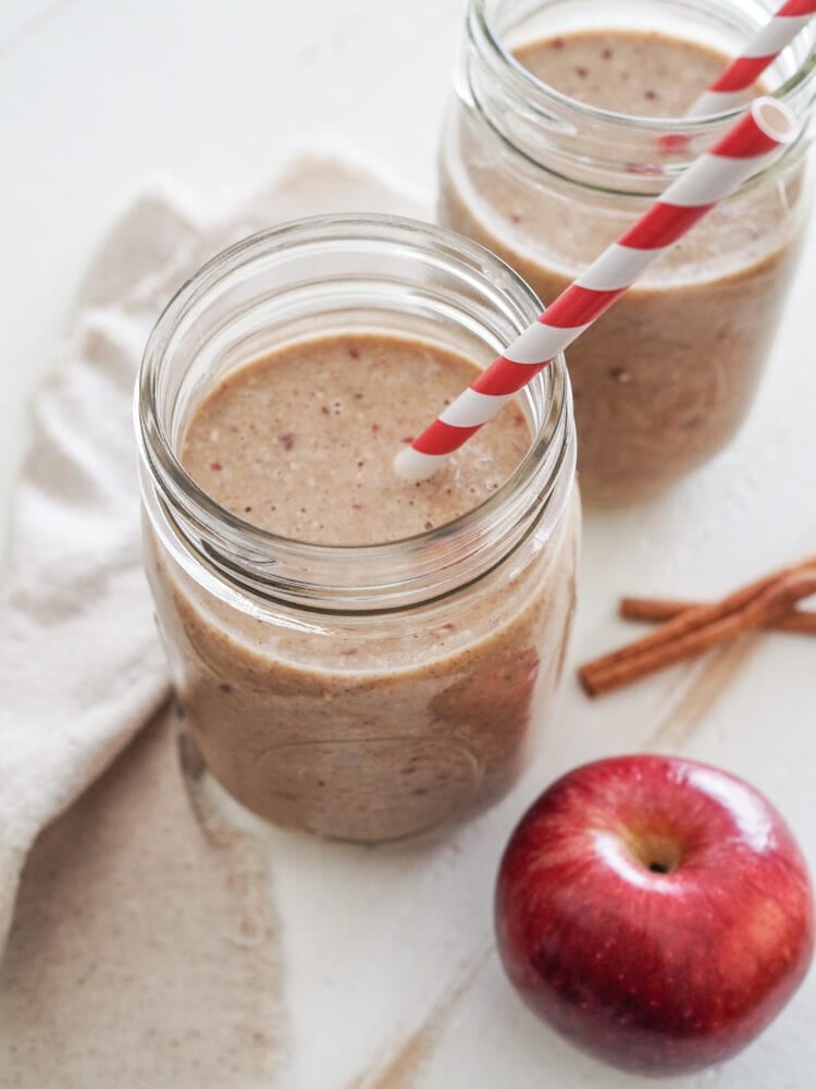 Two apple smoothies in mason jars with a red and white striped straw and a whole apple and cinnamon sticks to the side.
