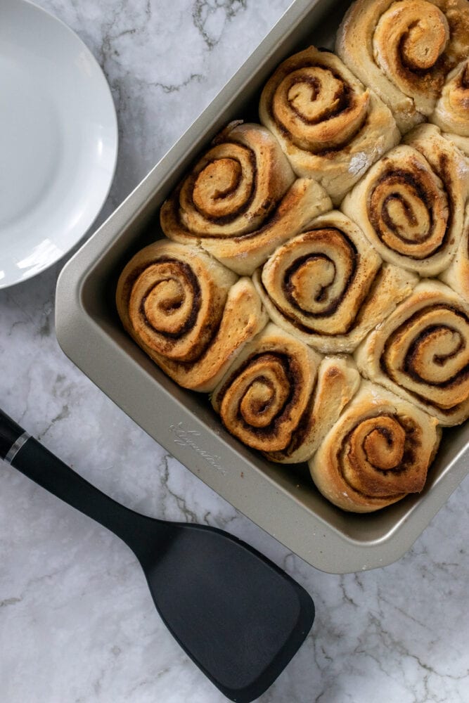 Overhead look into a metal baking pan with a dozen baked homemade cinnamon rolls, spatula and plate ready for serving.