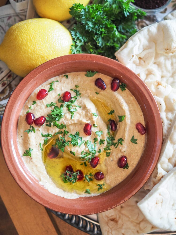 Baba ganoush, a roasted eggplant dip, in a terra cotta bowl garnished with pomegranate seeds, parsley, and olive oil. Shown with Naan bread, and lemons and parsley as decoration. 