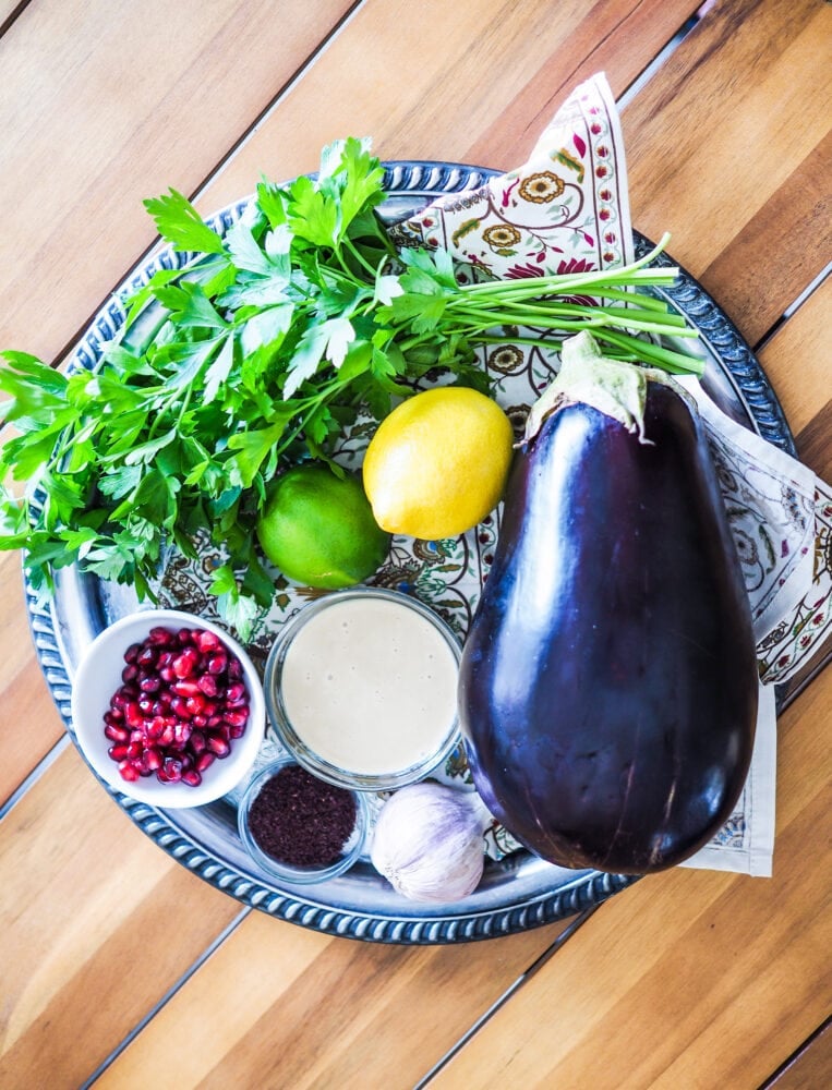 A platter showing some of the ingredients used to make baba ganoush including a whole eggplant, lemon, lime, tahini, garlic, and pomegranate, sumac, and fresh parsley.