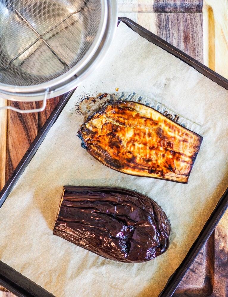 Roasted eggplant on a parchment lined cookie sheet. The skin of the eggplant is wrinkled and roasted, and one half of the eggplant is turned over flesh side up to show the browning from roasting it in the oven. There is a strainer and bowl off to the side prepped for removing the excess liquid and seeds from the eggplant.