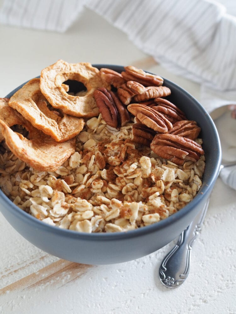 Bowl of microwave oatmeal on white table with dried apple slices, pecans, and cinnamon. 