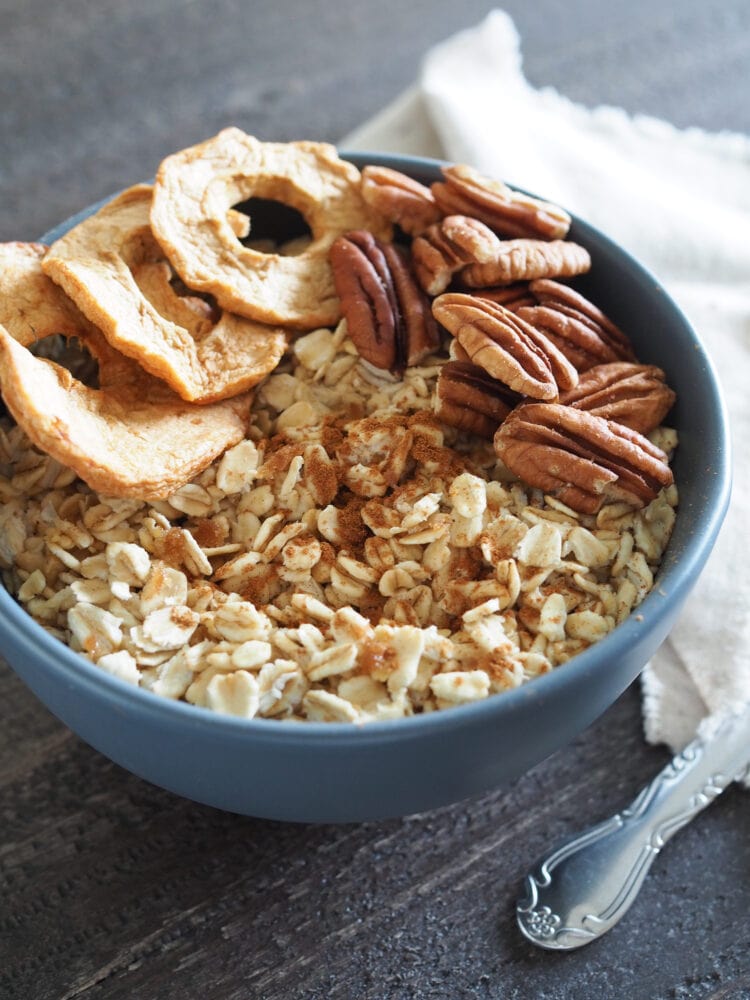 Bowl of microwave oatmeal on wood table with dried apple slices, pecans, and cinnamon. 