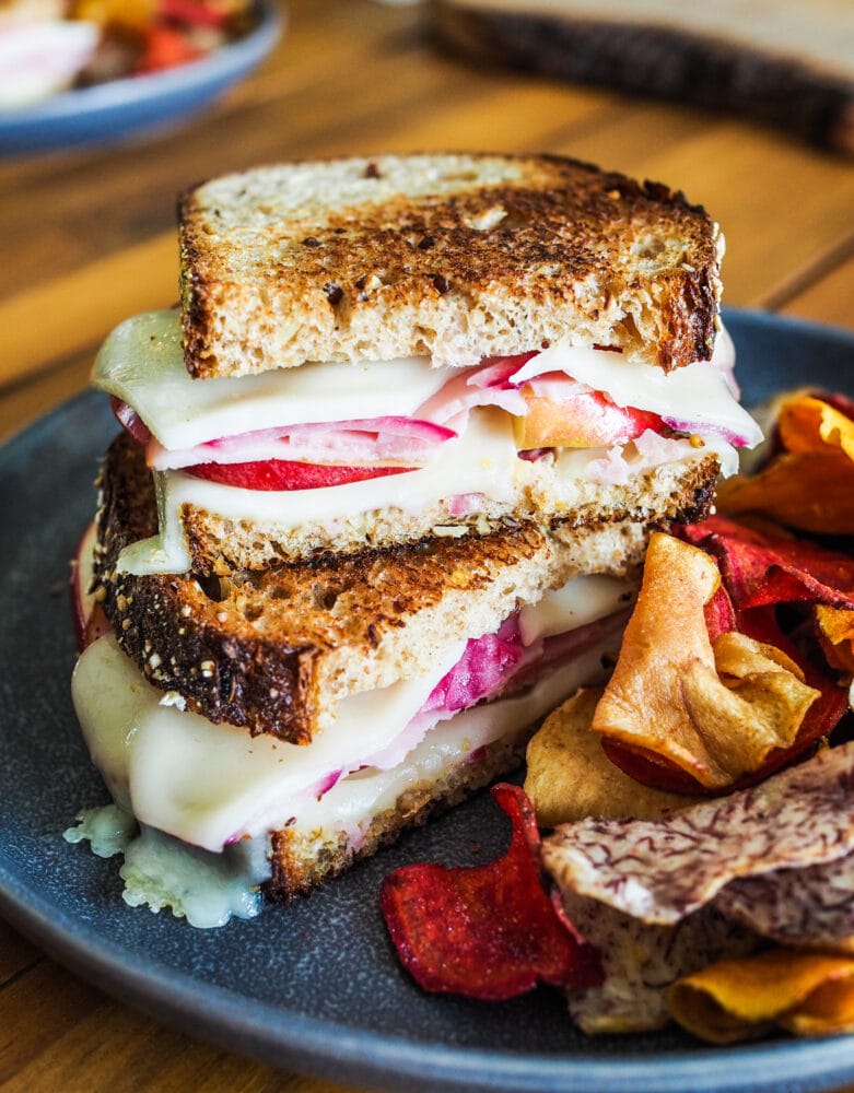 Stacked ham and cheese sandwiches with apples and pickled red onions served with orange and red root vegetable chips.