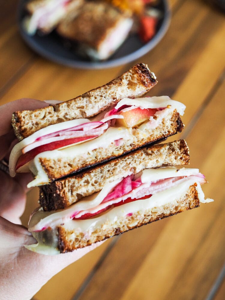 Hand holding two servings of ham and cheese sandwiches with apples and pickled red onions