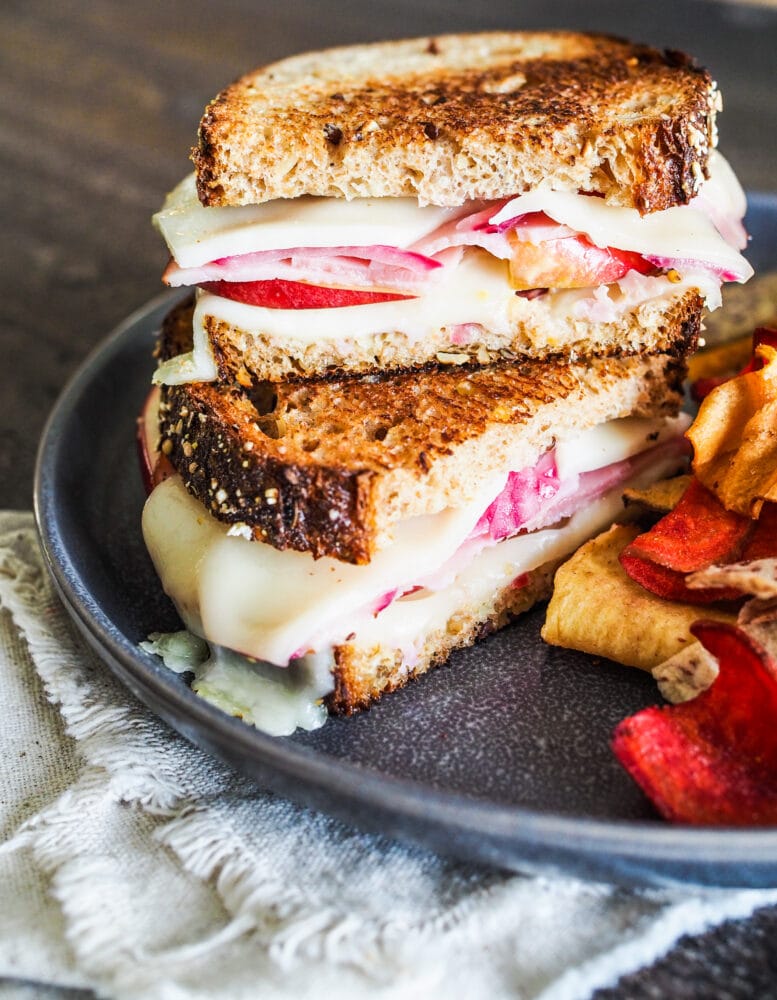 Stacked ham and cheese sandwiches with apples and pickled red onions served with orange and red root vegetable chips.