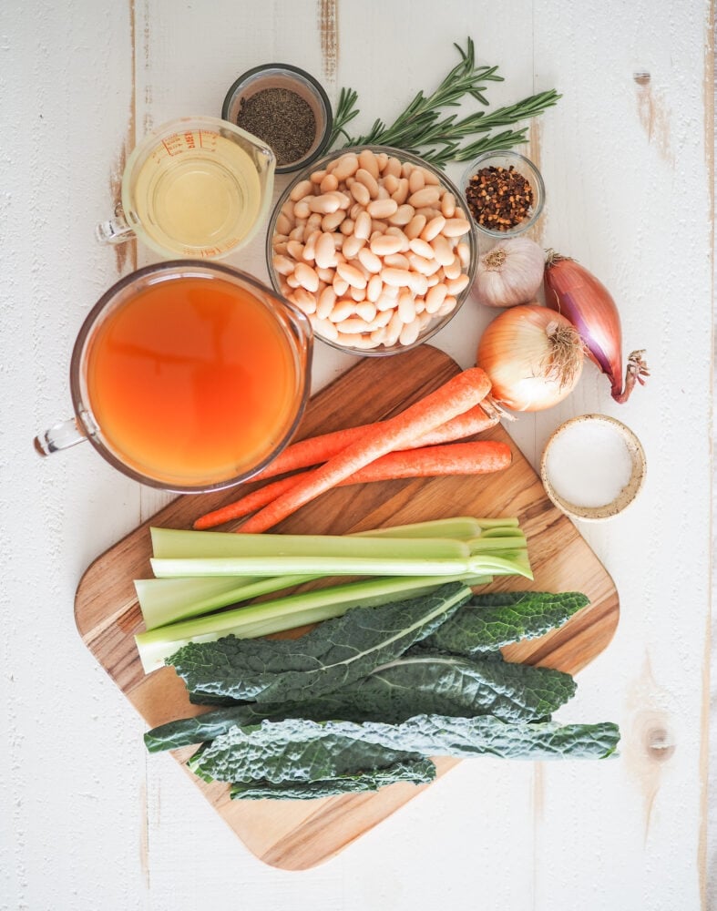 Overhead shot of ingredients for Tuscan white bean soup including: white wine, vegetable broth, cannellini beans, rosemary, red pepper flakes, garlic, shallot, yellow onion, carrots, celery, and kale.