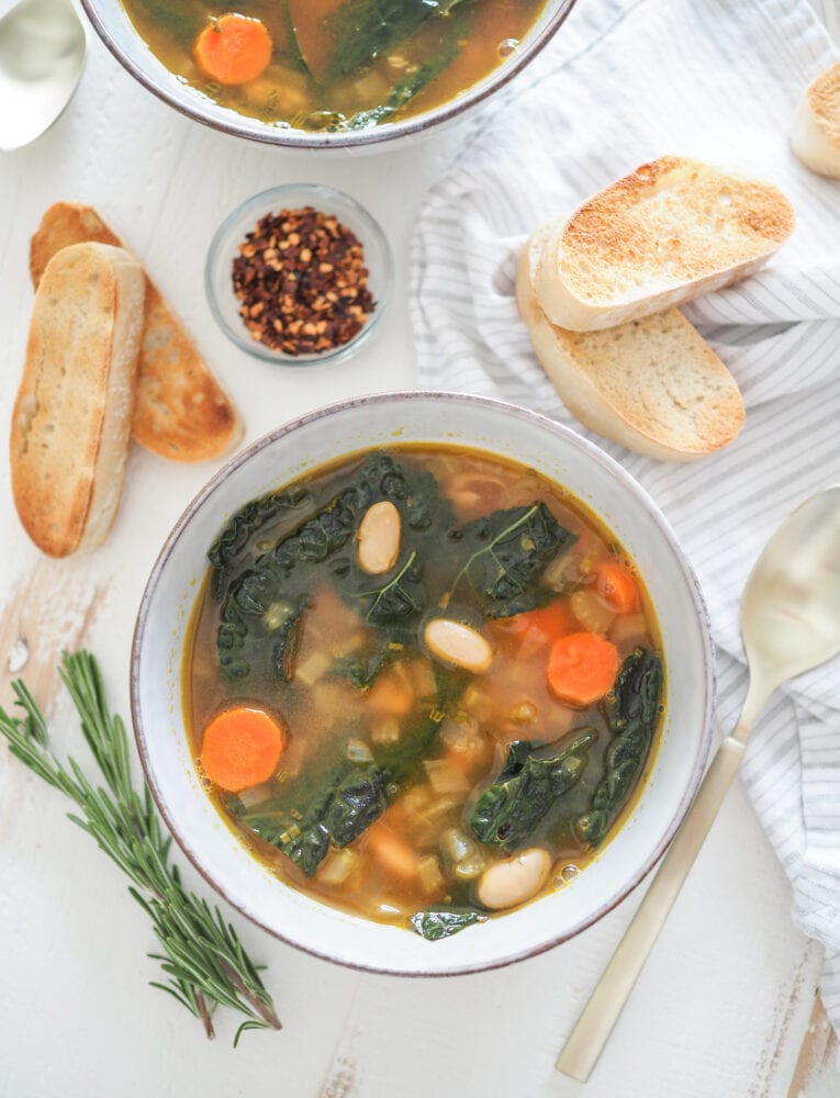 Two bowls of Tuscan white bean soup with a side of toasted ciabatta and small pinch bowl of red pepper flakes.