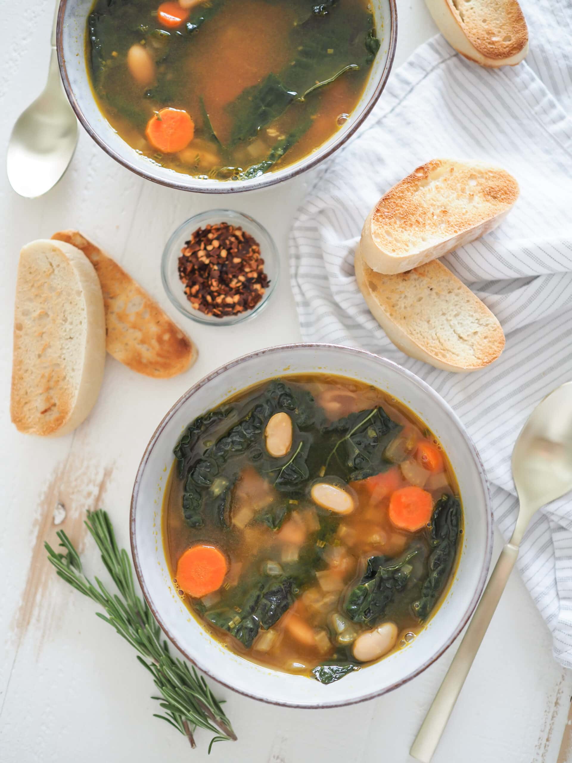 Two bowls of Tuscan white bean soup with a side of toasted ciabatta and small pinch bowl of red pepper flakes.