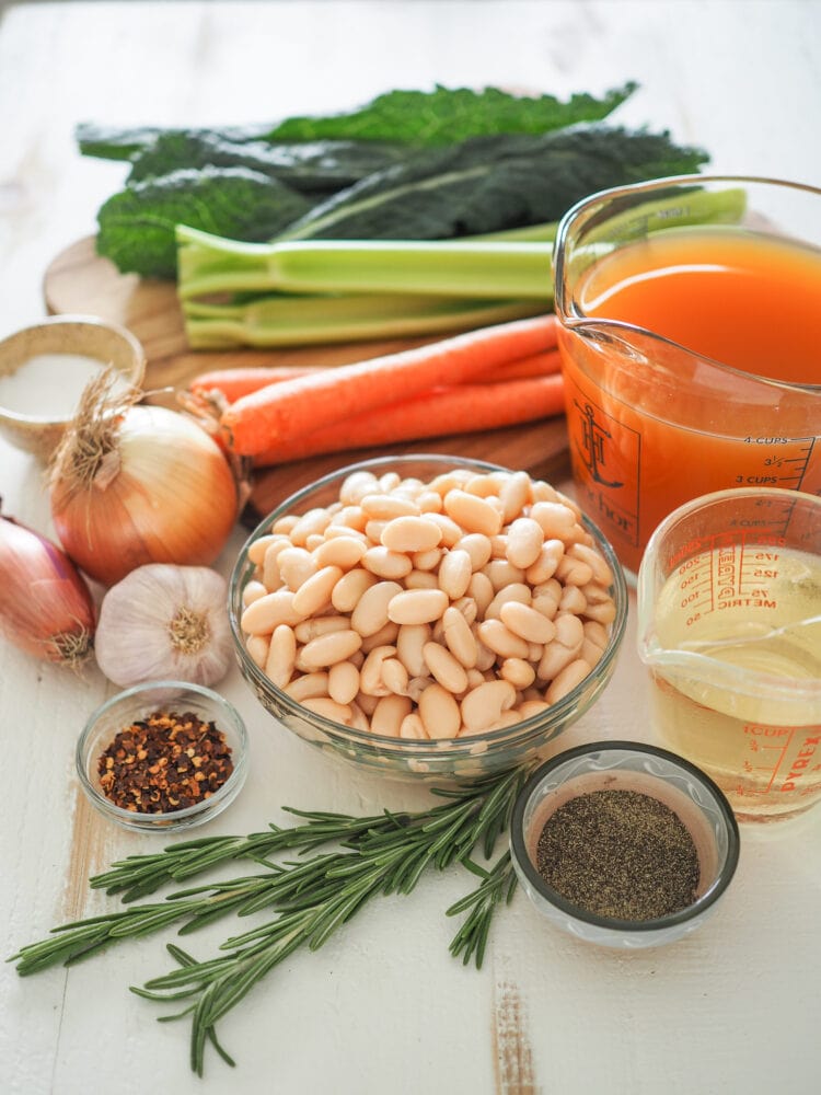 Side shot of ingredients for Tuscan white bean soup including: white wine, vegetable broth, cannellini beans, rosemary, red pepper flakes, garlic, shallot, yellow onion, carrots, celery, and kale.