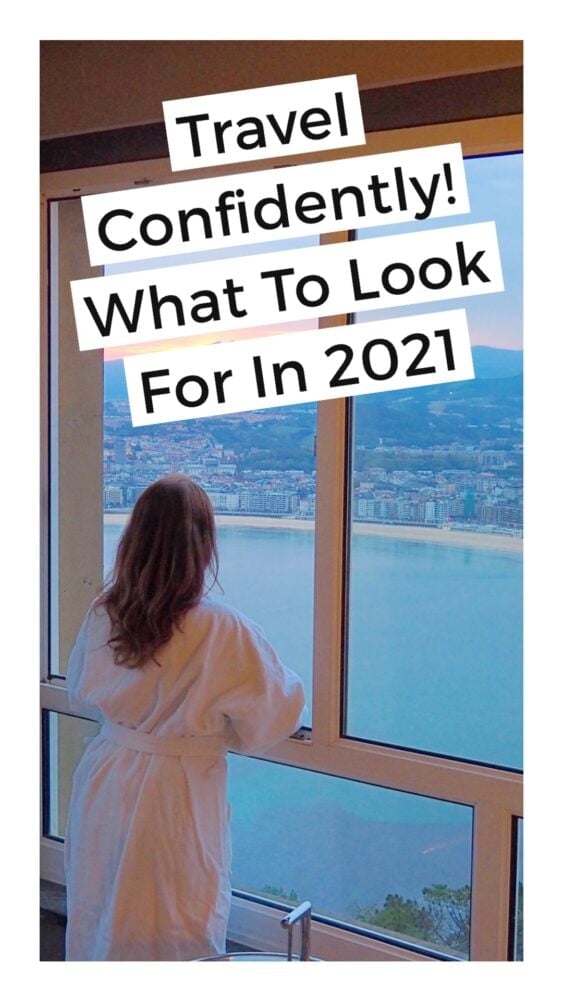 Girl wearing a robe looking outside of hotel window at a coastline with overlay text that reads, "Travel Confidently! What to look for in 2021"