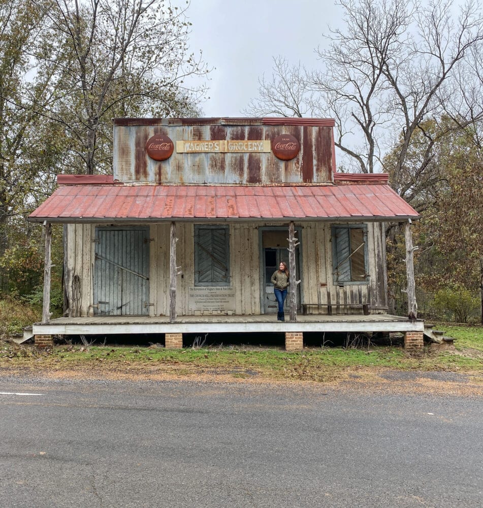 Rachelle standing on the porch of the old, rusty, wood historic building of Wagner's Grocery. There are light blue shutters closing up the old place and vintage CocaCola signs on the roof.