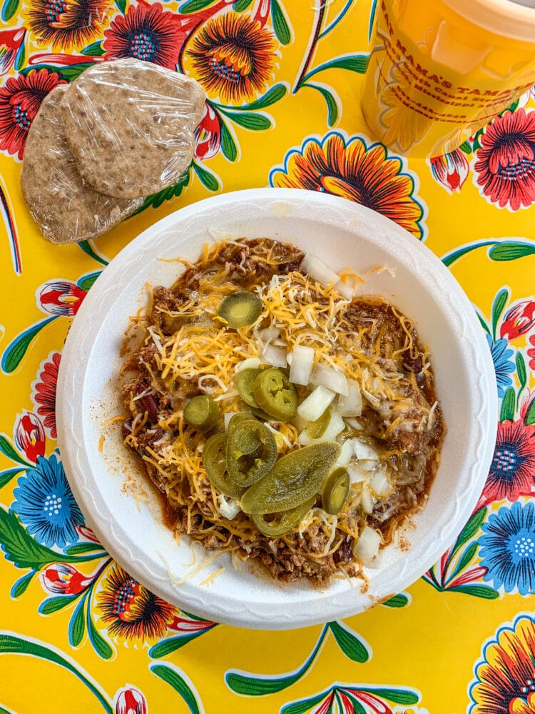 Overhead shot of tamales covered in sauce and cheese, pickled jalapeños, and chopped white onions, on a colorful yellow, blue, and red flowered table cloth. There are plastic wrapped pralines next to the plate (for dessert) and a yellow plastic cup filled with tea.