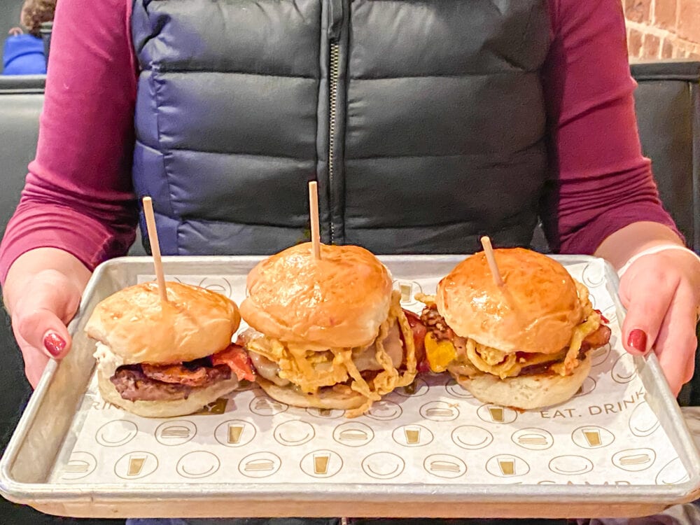 Rachelle holding a burger flight of three burgers on a small metal tray. 