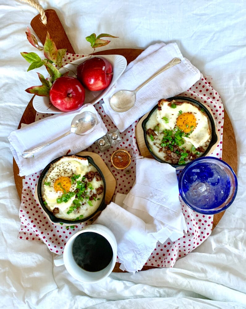 Overhead shot of a breakfast board in bed. There's a cup of coffee, apples, and small cast iron skillets with eggs and grits.