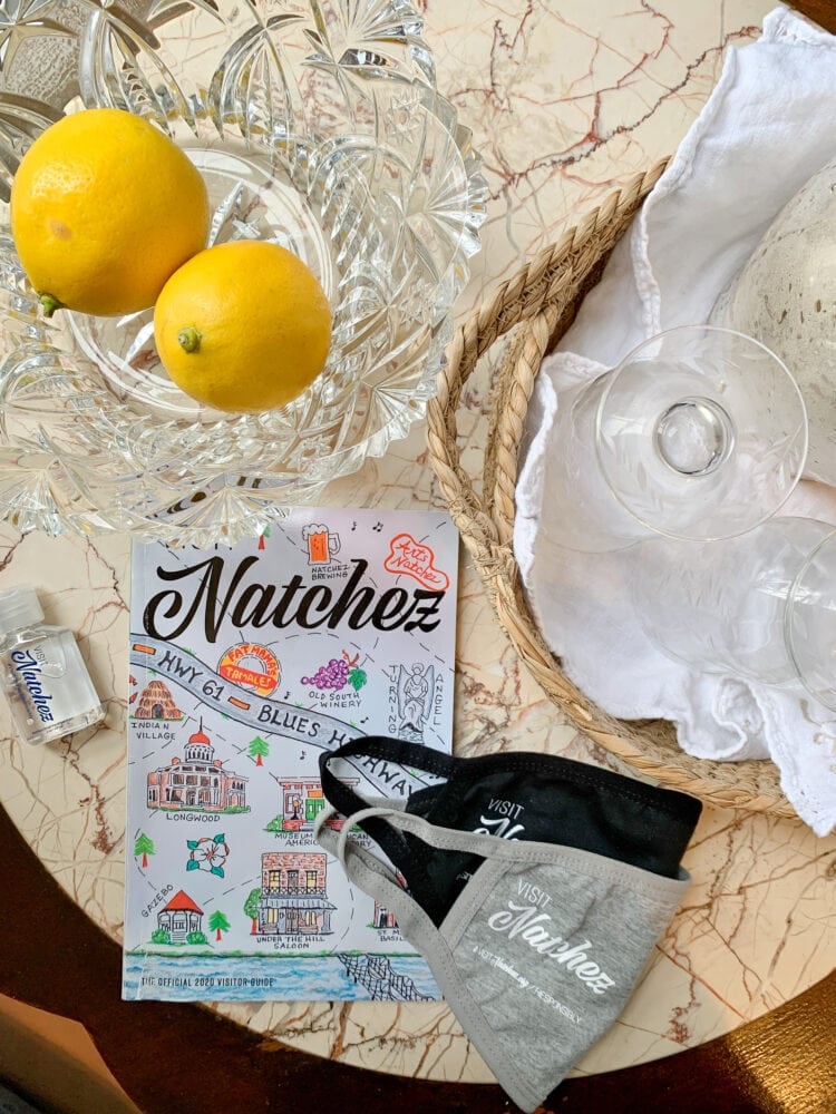 Natchez visitor's guide on a small table with a bowl of lemons, drinking glasses, hand sanitizer, and cloth face masks.