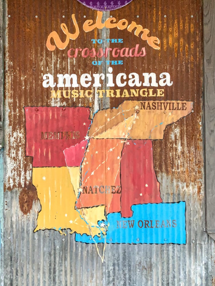 A rustic map on corrugated metal featuring Arkansas, Louisiana, Mississippi, Tennessee, and Alabama, showing the cities of Memphis, Nashville, and Natchez with stars. The sign says, "Welcome to the crossroads of the Americana music triangle."