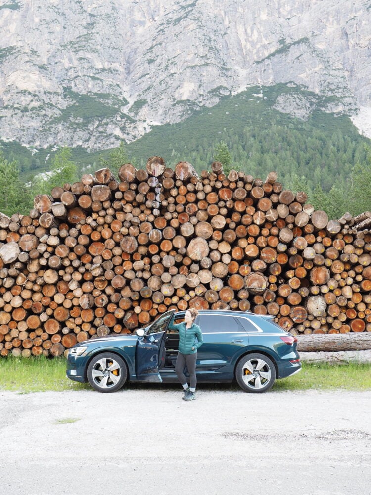 Rachelle wearing a puffer jacket and leggings, standing outside an SUV parked in front of stacked logs.