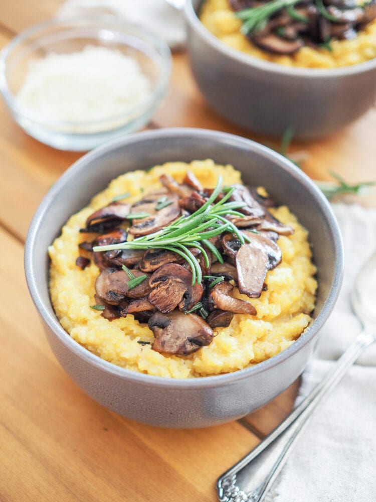 A wood dining table with a bowl of yellow cornmeal polenta topped with sautéed mushrooms and a fresh spring of rosemary. There's a bowl of Parmigiano-Reggiano cheese on the side.
