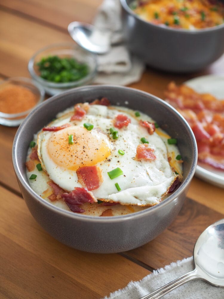 A bowl of grits topped with a fried egg, bacon bits, chives, and salt and pepper.