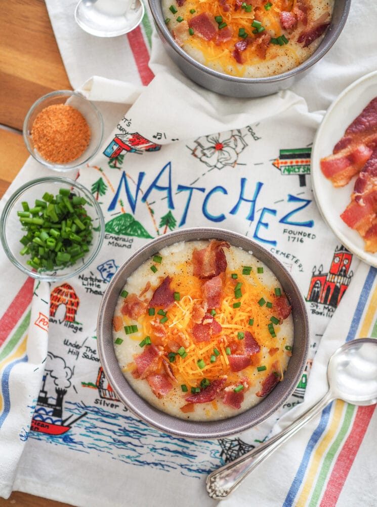 Overhead shot of a bowl of grits topped with cheddar cheese, bacon, and chives displayed on a kitchen towel that says "Natchez."