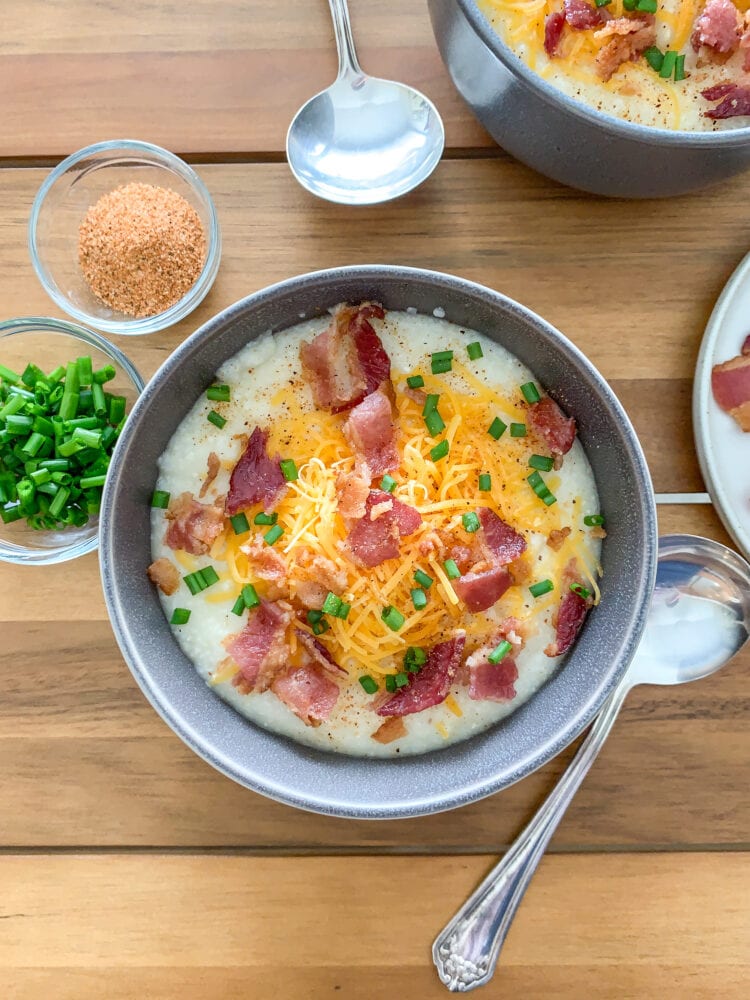 Overhead shot of a bowl of grits topped with cheddar cheese, bacon, and chives with pinch bowls of extra chives and seasoning on the side.